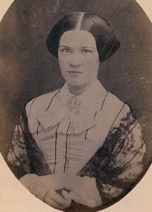 Black and white photograph of a woman from the waist up in a white dress with narrow black stripes and a black lace shawl.