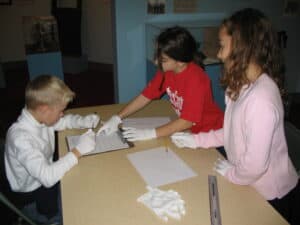 A boy and two girls around a table wearing white gloves and writing.