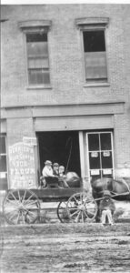 A horse drawn wagon in front of a brick storefront.  Three people are sitting in the wagon and one person is sitting on the ground along side the horse.
