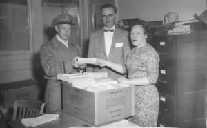 two men and a woman standing behind a box on a desk in an office