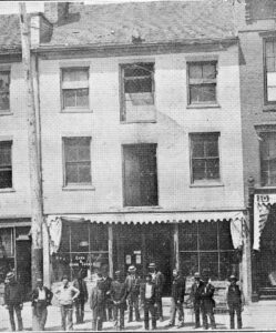 A group of men standing outside a 3-storey building with glass plate store windows and an awning on the first floor.