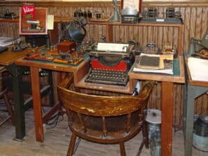 A small wooden desk with a typewriter and telegraph machines on it and and banker's chair in front of it.