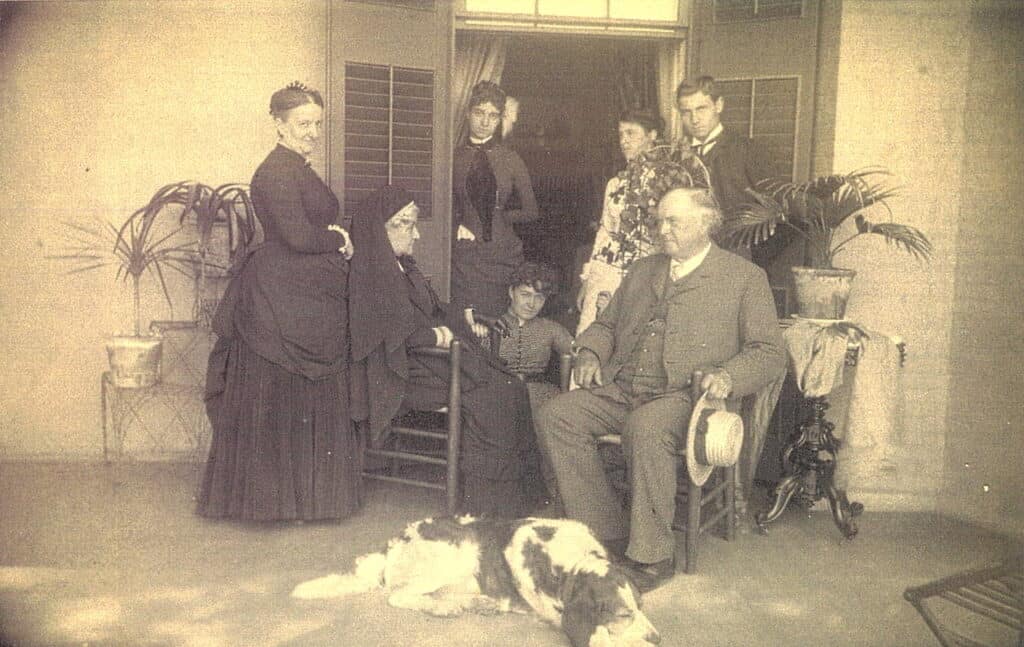 A group of people in front of a door with a dog at the feet of an older man sitting in a chair and holding a straw hat.