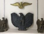 Shelf display of four metal eagles. The center one is large and black iron with wings spread Hanging above it is a small gold one and it is flanked by twin brass ones. and one on either side.