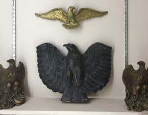 Shelf display of four metal eagles: at center a large, black iron one with wings spread, flanked by twin brass eagles and a small gold one hainging above.