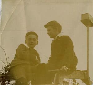 Two boys in short pants, caps and jackets sitting on top of an arbor.
