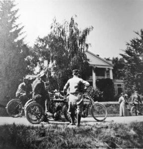 group of men with motor cycles and two women with a man on a bicycle are in front of Maple Hill