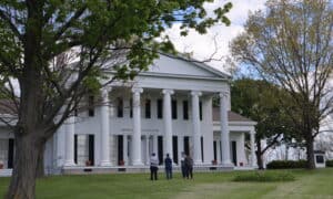 A group of people standing on a lawn in front of a large, white, 6-columned house.