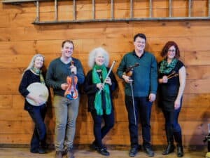 A group of men and women standing in front of a wooden wall. Each holding a different instrument: drum, fiddle, flute, fiddle and dulicmer hammers.