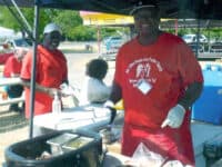 A smiling black man in a red and white Mount Olive Missionary Baptist Church t-shirt, red apron, and black ball cap prepareing food in a booth outdoors. Other people stand or sit in the background.