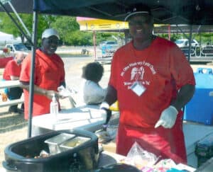 A smiling black man in a red and white Mount Olive Missionary Baptist Church t-shirt, red apron, and black ball cap prepareing food in a booth outdoors. Other people stand or sit in the background.