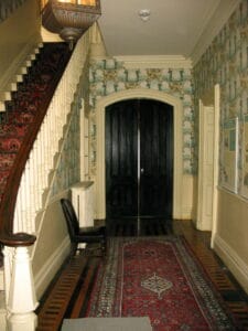 View into a large entryway with a staircase to the left, double wooden doors at the end, oriental carpets on the floor, and green wallpaper.