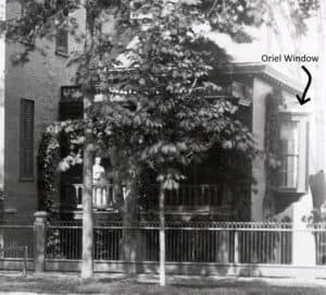 A woman standing on the porch of a house mostly hidden by foilage. The porch has a window on the end that butts out and is labeled oriel window.