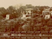 An old photo of a hillside with houses on it terraced above a train track and water.