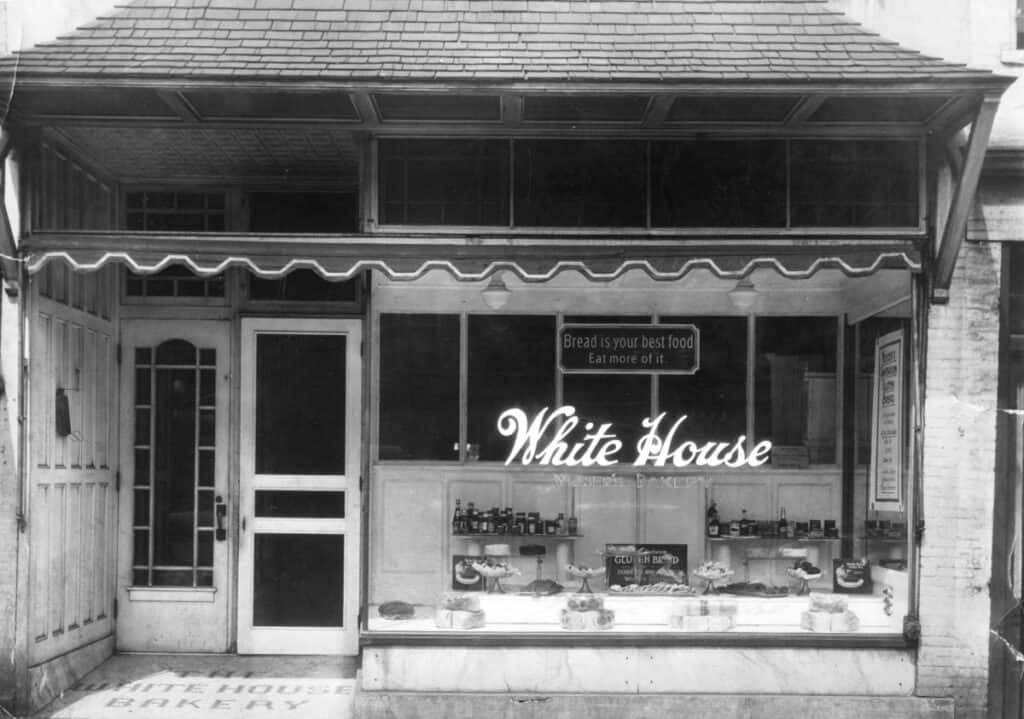 A store front with a glass display window and the label White House Weiser's Bakery. In the window are a variety of baked goods, bottles and jars. A sign says Bread is Your Best Food Eat More of It.