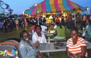 Three young black women sitting around a picnic table at a festival at twilight. In the background are a crowd of people, amusement park rides and striped tents.