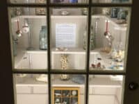 View of a cabinet with 4 levels of shelves with a display case and glass jars full of seashells and books and a scatter of seaglass on the floor.