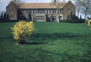 View across a wide green lawn with a forsythia bush on it towards a large brick and stone two-story building with a flagpole out front.