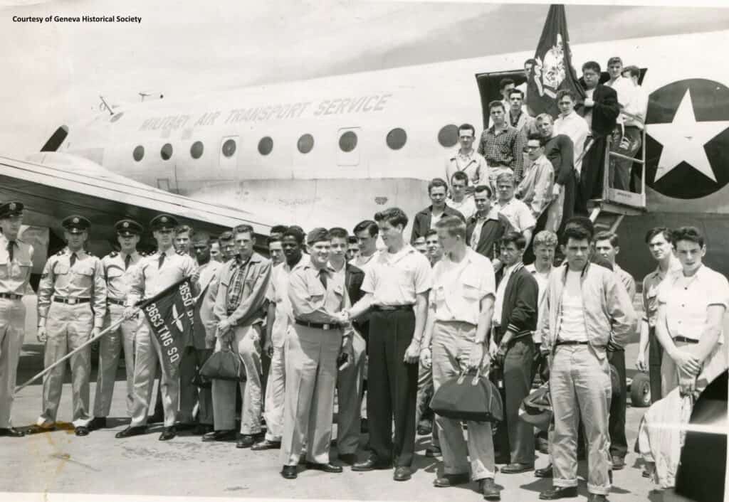 A large group of very young men in civilian clothes standing with men in military uniforms front of a plane labeled Military Air Transport Service.