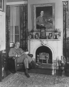 A balding middle-aged man with glasses in a 3-piece suit sits reading in a a leather armchair by a marble fireplace over which hangs a painting of the man.