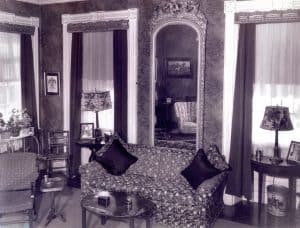 A living room with heavy draperies and cornices on the windows and furnished with a sofa, chairs, tables, and an antique gilded ceiling-height mirror.