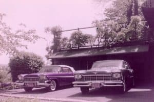 A faded color photograph of 1950s cars parked side-by-side in front of a garage.