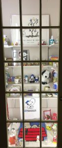 View of a cabinet with 3 levels of shelves with Snoopy-themed prints, mugs, hooked art, stuffed animals and figurines on them.