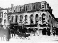 People and a horse-drawn sleigh standing around the street outside a badly fire-damaged building of three stories and a mansard roof.
