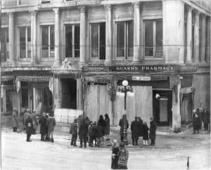 People standing outside a commercial building with classical columns that is boarded up and damaged.