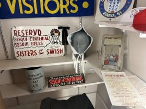 Three shelves in a cabinet with a display of signs, hats and a other items marking Geneva's sesquicentennial in 1957.