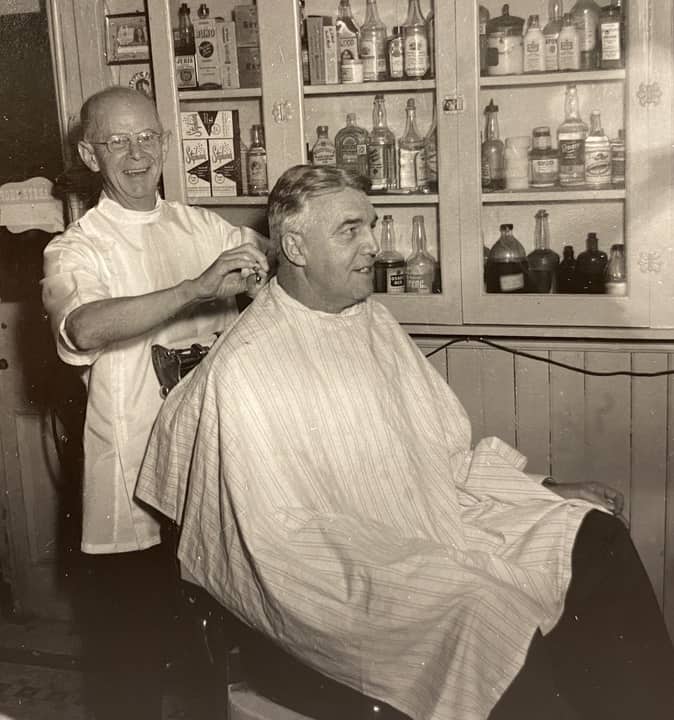 A man sits in a chair with a white cape over his clothes while a balding man with glasses in a white smock cuts his hair.