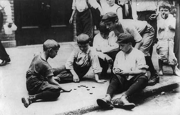 A group of boys in shirts, knickers and stockings playing checkers on a city sidewalk.