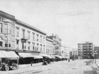 View down a street of commercial buildings, many with awnings and 1920s Model Ts parked on the street.