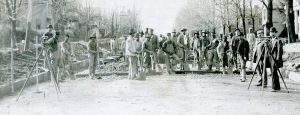 A large group of black and white men in suits and overalls stand in the middle of a road among piles of rock, shovels, wheel barrows, and several surveyors' tools.