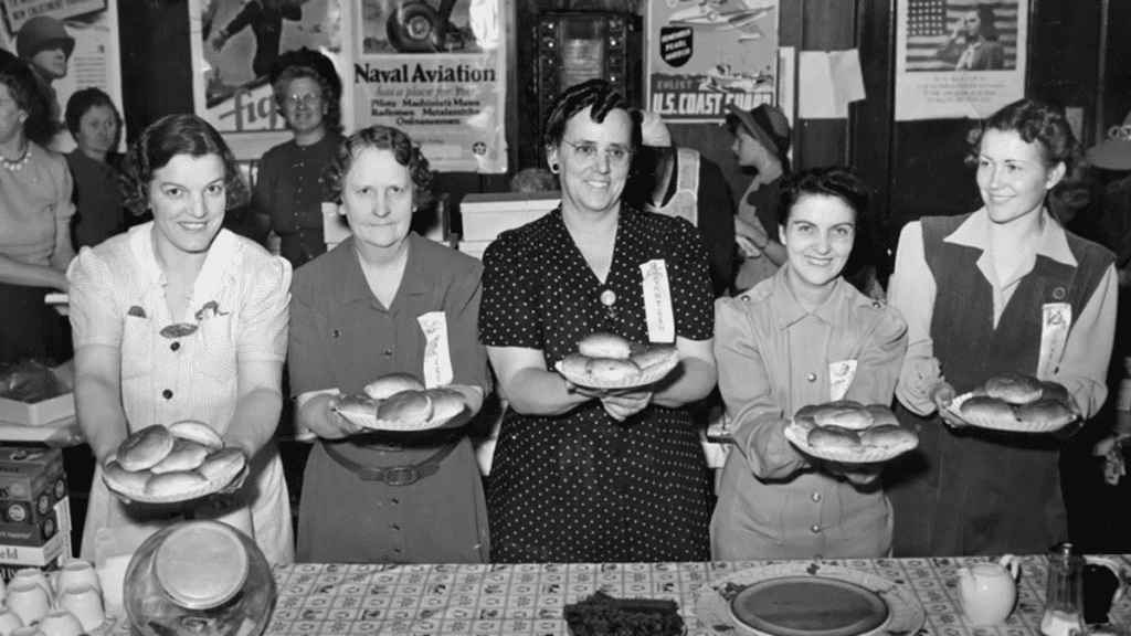 Five women in 1940s dresses standing behind a table and holding out plates of rolls.