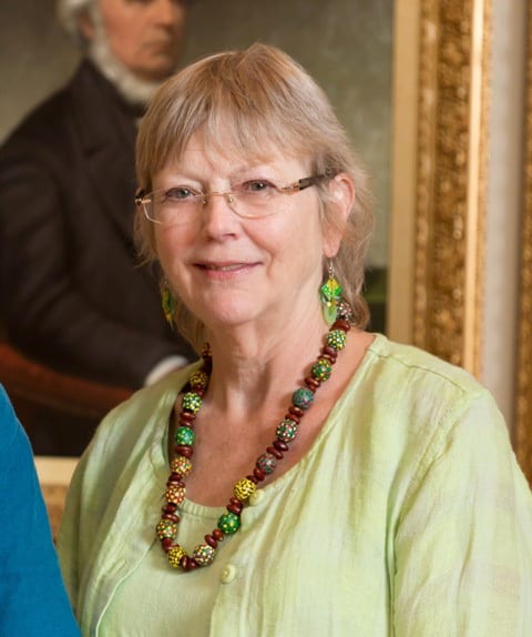 Headshot of a middle-aged blond woman with wire rimmed glasses, a light green shirt and a chuncky brown and green bead necklace and earrings.