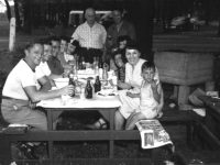 A group of men, women and children sitting and standing around a picnic table covered with bottles and dishes.