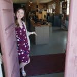 A young woman in a maroon and white flower print dress stands at the open door of a room with a long lab table with instruments on it.