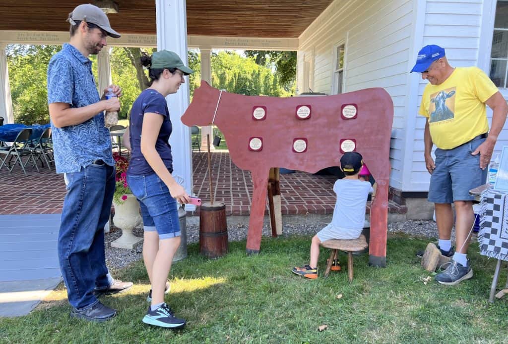 A couple and an older man watch a little boy sit and "milk" a plywood cow.