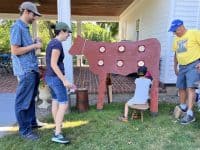A couple and an older man watch a little boy sit and "milk" a plywood cow.