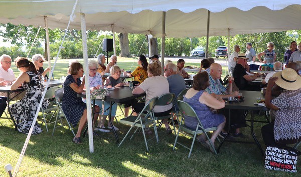 A group of people sitting at tables under a tent on grass on a sunny evening.