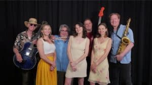 Four middle-aged men each holding a guitar, bass, drumsticks or sax, posing with three young women in front of a black curtain.