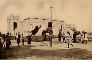 Six girls on an early playground swinging around a central pole from ladders hanging from its top.