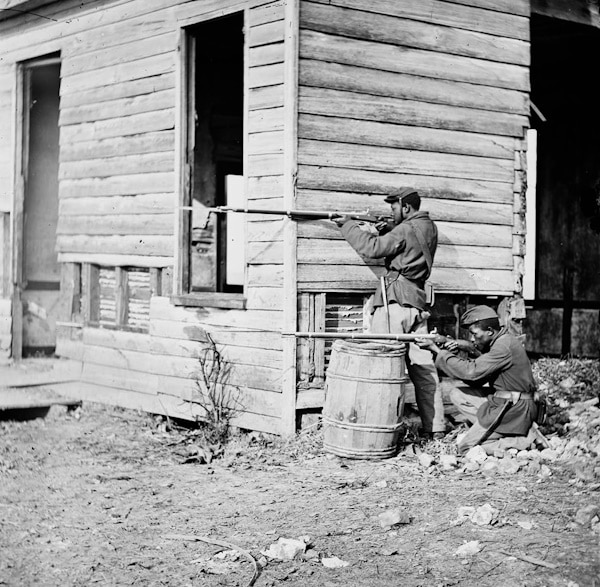 Two black Union soldiers aiming rifles from behind a building and a barrel.