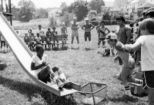 A large group of children and young men stand around a playground slide while a boy slides down it holding something.
