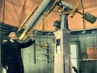 A man in a black suit standing with his eye to an old-fashioned telescope.