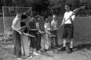 A young man stands on a field with six boys who are all holding lacrosse sticks in front of a net.