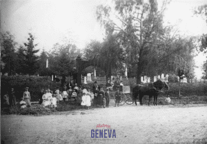 Large group of people and a horse-drawn buggy are gathered in front of a cemetery entrance.