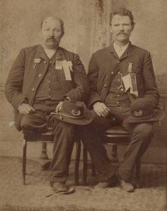 Two men sitting with military hats on their laps. One man is missing a leg.