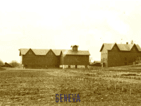 Black and white view across bare fields towards five buildings including an Italianate house and several barns.